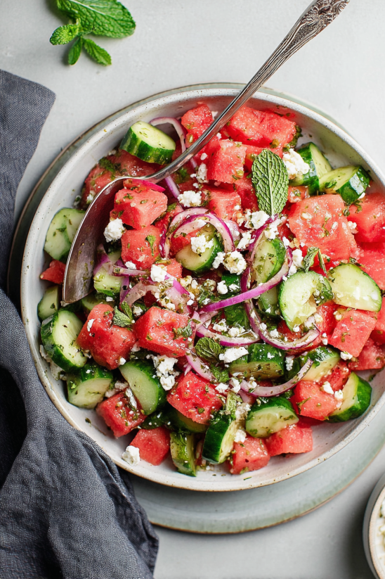 A bowl of fruit salad with watermelon, cucumbers, and onions.