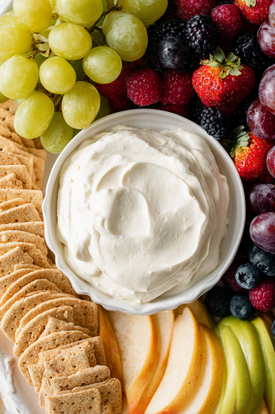 A bowl of cream cheese sits on a table with a variety of fruits.