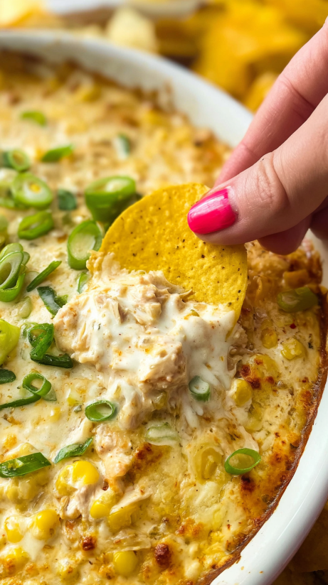 A person is holding a tortilla chip over a baked corn dip.