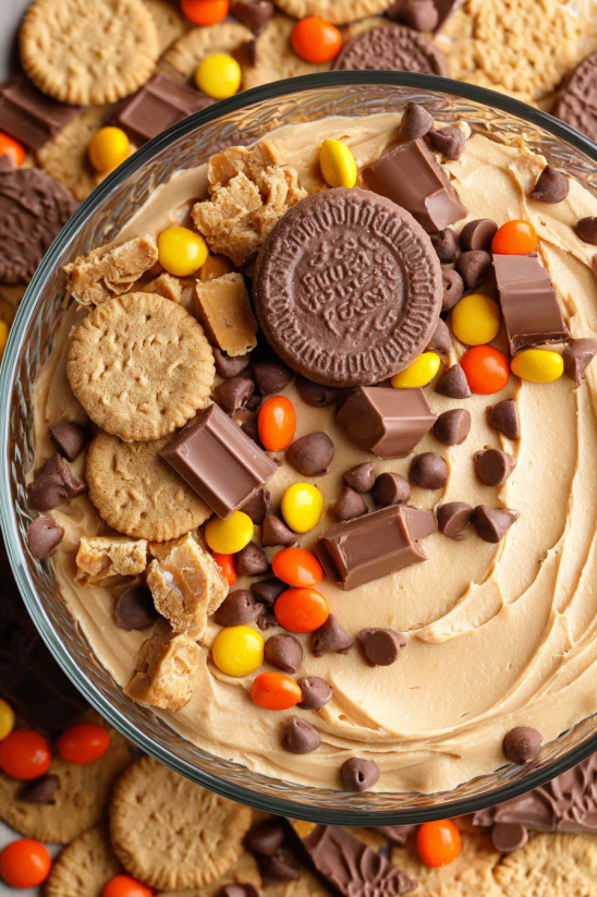 A bowl of peanut butter cheesecake dip with chocolate chips and candy.